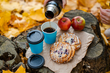 Autumn picnic concept - female hand pouring hot coffee into cup on tree stump with pastries and apples among colorful leaves