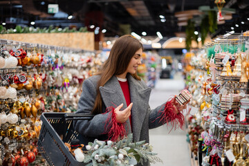 Young woman choosing Christmas ornaments in a festive store aisle, holding decorations while shopping with a basket.