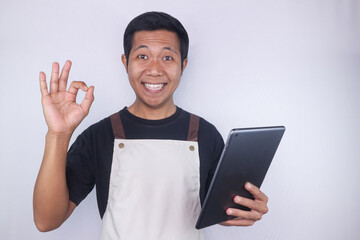 Smiling young Asian man barista barman employee wearing apron hold digital tablet showing ok gesture