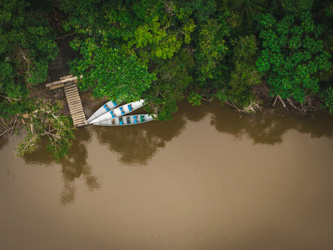 Aerial view of three boats resting by a wooden pier on the murky riverbank, framed by the vibrant green canopy of the Amazon Rainforest, Tipishca, Sucumbios, Ecuador.