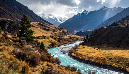 A vibrant turquoise river flows through a valley with golden grass and sparse trees, leading to majestic snow-capped mountains under a cloudy sky.