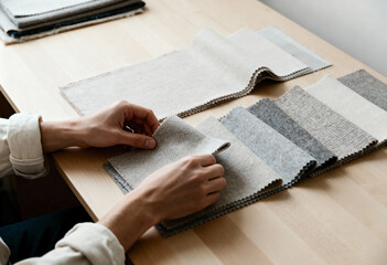Designer's hands choosing from neutral fabric swatches on a wooden table. Person examining textile samples with different textures for an interior design project