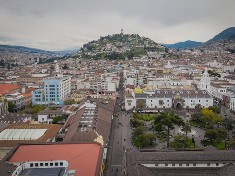 Aerial view of a sprawling cityscape unfolds beneath a cloudy sky, leading towards the iconic Panecillo hill topped with a statue, Quito, Ecuador.