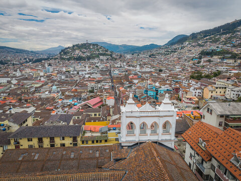 Aerial view of a sprawling cityscape under a cloudy sky, rooftops cascading towards the distant Panecillo hill and mountains, Quito, Ecuador.
