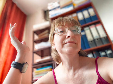 Mature blonde woman sharing her blogging experience during a casual selfie at home with bookshelves in the background