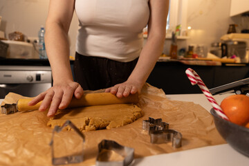 A person rolls out gingerbread dough on a kitchen counter filled with festive items. Various cookie cutters and a candy cane add to the Christmas spirit as they enjoy baking.
