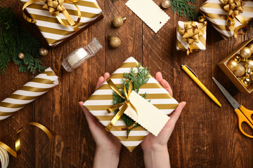 Christmas present packaging. Hands holding a ready-made gift wrapped in white and gold striped paper with a blank tag for your text.