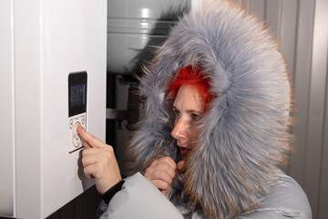 A woman wearing a winter coat with a furry hood pushes a button on a white thermostat. She is...
