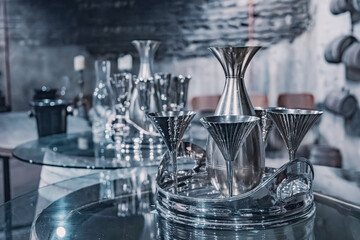 Silver drinkware set with decanter and cocktail glasses on a glass table within a dimly lit, luxurious lounge setting