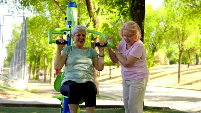 Two cheerful senior women enjoying an active lifestyle, exercising on outdoor gym equipment in a sunny park. One woman uses a shoulder press machine while her friend provides motivation and support