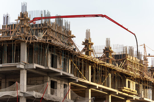 A detailed view of a construction site with a red concrete pump boom extending over the formwork and rebar of a new building. The process of pouring concrete is underway.