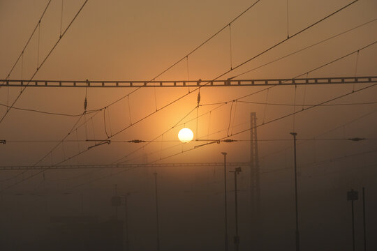 A dramatic foggy sunrise over a railway line. Silhouettes of overhead power cables crisscross against the golden sun and orange sky in a misty atmosphere