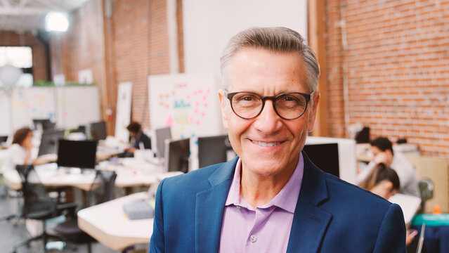 Portrait Of Mature Businessman In Modern Open Plan Office With Business Team Working In Background