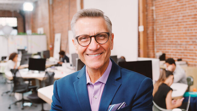 Portrait Of Mature Businessman In Modern Open Plan Office With Business Team Working In Background