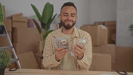 Man counting us dollar cash with hands at wooden table among moving boxes and houseplants in building; financial success satisfaction.