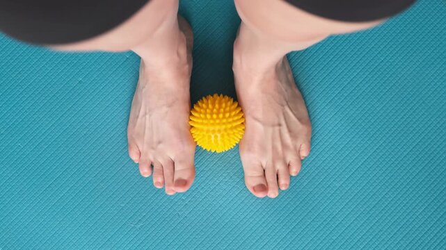 Female feet are doing foot exercises picking up the prickly massage ball rolling from heel to toe standing on the yoga mat. Training to correct and relax the foot fascia and muscles. 4k footage.
