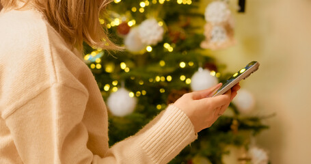 Cozy clothed woman browsing smartphone near softly lit Christmas tree, immersing in festive home...