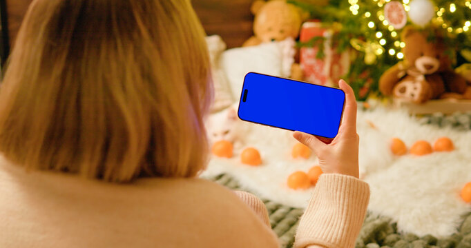 Winter holiday scene featuring female relaxing near decorated Christmas tree, holding smartphone with blue screen mockup for digital content creation