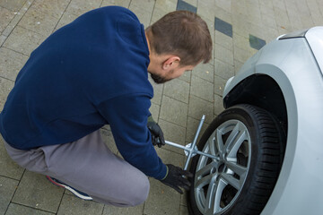 Man changing car tire using electric drill and jack on city sidewalk. DIY auto repair, maintenance, seasonal tire change, mechanical work, vehicle service, urban life.