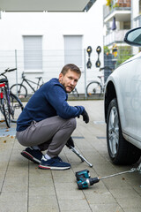 Man changing car tire using electric drill and jack on city sidewalk. DIY auto repair, maintenance, seasonal tire change, mechanical work, vehicle service, urban life.