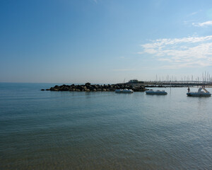 Obraz premium A serene, wide-shot photograph of a calm, blue sea and sky, featuring a short rock breakwater in the middle ground and small boats pedal boats anchored near the shore.