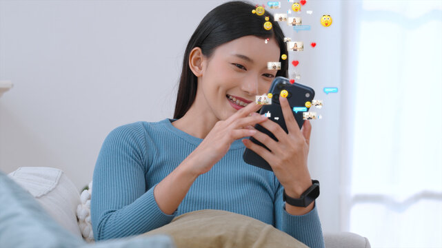 Smiling young woman sitting on a couch using smartphone with floating social media icons and emoji reactions around her.