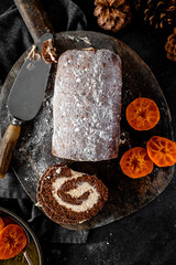 Homemade chocolate roulade filled with vanilla cream, dusted with icing sugar, and served alongside glossy candied orange slices. Presented on a rustic wooden board with pinecones and dark textured ba
