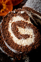Homemade chocolate roulade filled with vanilla cream, dusted with icing sugar, and served alongside glossy candied orange slices. Presented on a rustic wooden board with pinecones and dark textured ba