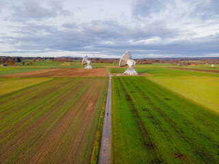Aerial view of two satellite dishes standing tall in a field of green and brown, under a cloudy sky, Hofstatterweg 1, Raisting, Bayern, Germany.