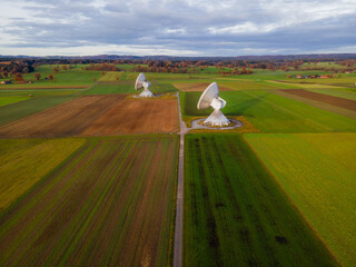 Aerial view of two massive satellite dishes standing as sentinels amidst a patchwork of green and brown fields under a cloudy sky, Raisting, Bayern, Germany.