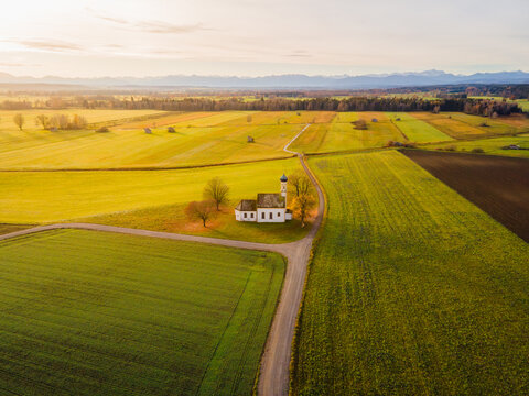 Aerial view of a quaint white church standing amidst a patchwork of golden and green fields under a serene sky, Raisting, Bayern, Germany.