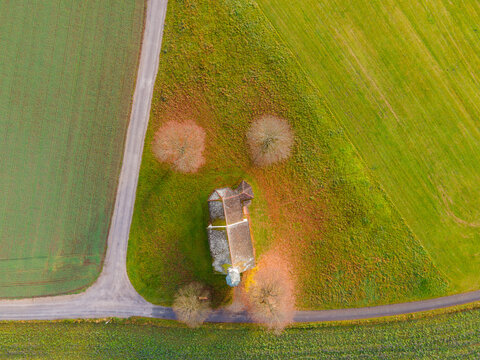 Aerial view of a quaint chapel nestled amid vibrant green fields, framed by bare trees and winding pathways, Raisting, Bayern, Germany.
