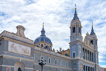 The Almudena Cathedral in Madrid, showcasing its neo-Romanesque exterior, with twin bell towers, a central dome, and a large stone staircase under a dramatic blue and white sky.