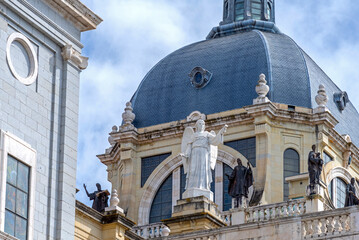 A close-up view of the Almudena Cathedral dome and its lower drum, featuring white and dark bronze statues atop an ornate balustrade against a bright, cloudy sky