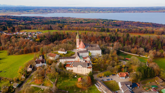 Aerial view of the Andechs Abbey standing majestically amidst a sea of golden trees, with the tranquil Ammersee lake shimmering in the distance, Andechs, Bayern, Germany.