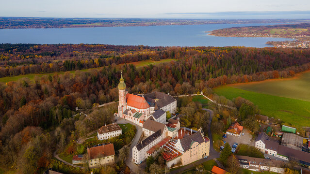 Aerial view of the Andechs Abbey with its red roofs and white walls nestled amidst the vibrant autumn foliage overlooking the distant lake, Andechs, Bayern, Germany.
