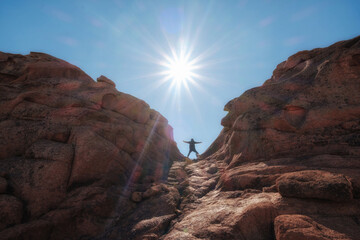 A man stands on the edge of a crevice between the red rocks of the Bektau Ata tract in Kazakhstan against the background of the bright sun, symbolizing the achievement of goals and adventures