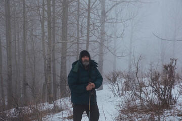 A bearded man in a warm jacket and hat stands with a staff on a snow-covered path in a misty winter forest, surrounded by bare trees.