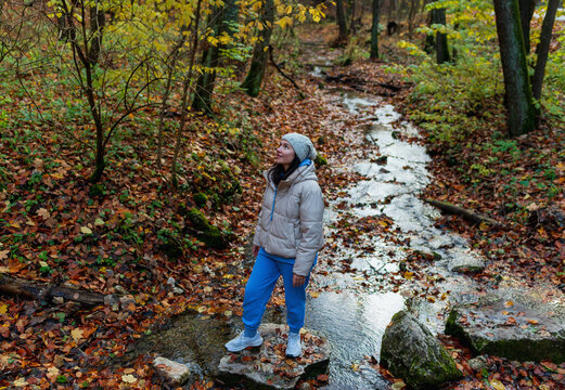 A lone woman stands on a stone in a shallow autumn stream, surrounded by fallen leaves and warm forest colors. The moment captures peaceful outdoor exploration, seasonal mood, and connection with natu