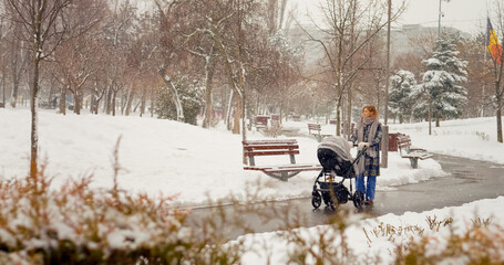 A young mother strolls through a peaceful park while snow falls gently around her. The serene winter landscape provides a beautiful backdrop for their outing.