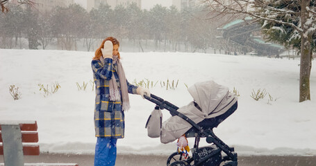 In a snowy park, a mother struggles with a headache while pushing her baby stroller along the path. The white landscape creates a serene yet challenging atmosphere for her stroll.