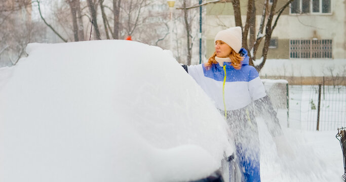 In a challenging winter setting, a woman braves the cold to remove heavy snow from her car, demonstrating determination as she navigates the frosty conditions outdoors.