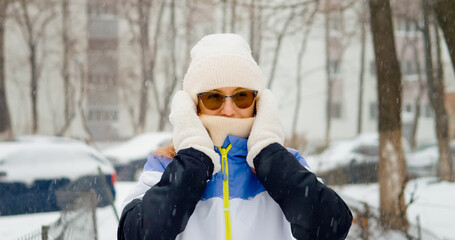A woman in winter attire stands outside in the snow, adjusting her hat while surrounded by a picturesque snowy landscape. It's a cold day filled with snowflakes.