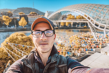 Man smiling, taking a self portrait while traveling, with the bridge and Kura river visible in the background, Tbilisi © EdNurg