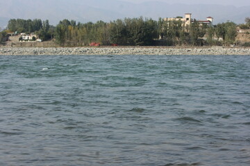 A wide view of a flowing river with prominent ripples and submerged stones in the foreground, bordered by a wide gravel bank and a distant line of trees and houses
