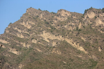 A steep, rugged mountain face with exposed layers of light-colored rock interspersed with patches...