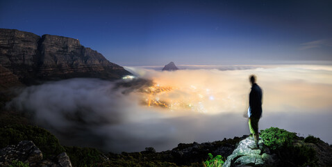 View of a solitary figure stands atop rocky terrain, gazing over a city shrouded in a thick blanket of fog illuminated with golden light, Cape Town, Western Cape, South Africa.