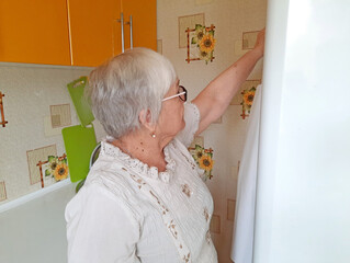 An elderly woman with glasses and gray hair stands in the kitchen, reaching for her apron.
