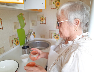 An elderly woman prepares pancakes in the kitchen, holding small ceramic pots in her hands.