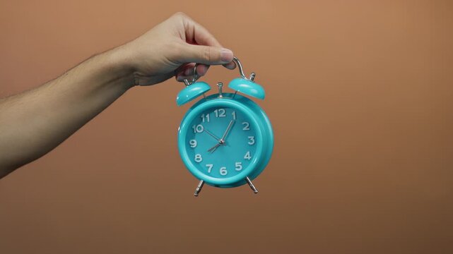 Man holding turquoise clock with brown wall background, emphasizing time management and punctuality with isolated hand in vibrant setting.